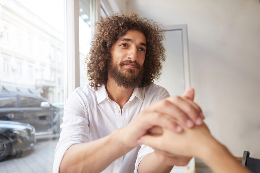 Young attractive curly man with beard and brown kind eyes sitting by the window, holding female hand and looking at her dearly, wearing white shirt